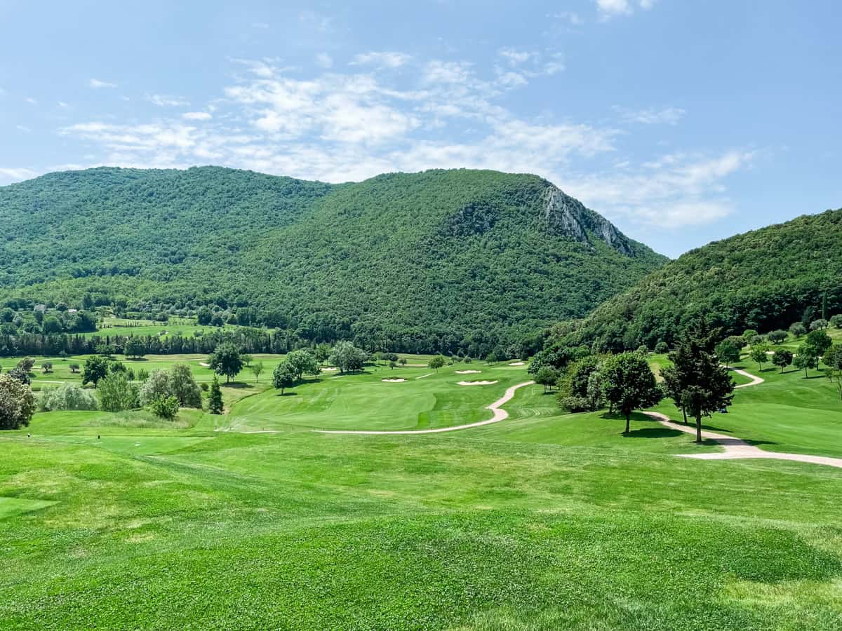 Wide view of a lush valley golf course surrounded by steep, tree-covered mountains, with rolling fairways, scattered bunkers, and winding cart paths under a bright blue sky with light clouds.