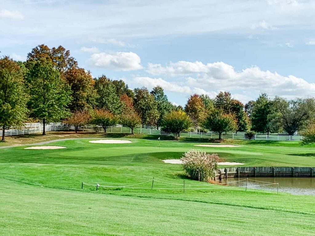 Golf green with multiple sand bunkers and a small water hazard, surrounded by trees with autumn foliage and a white fence under a partly cloudy sky