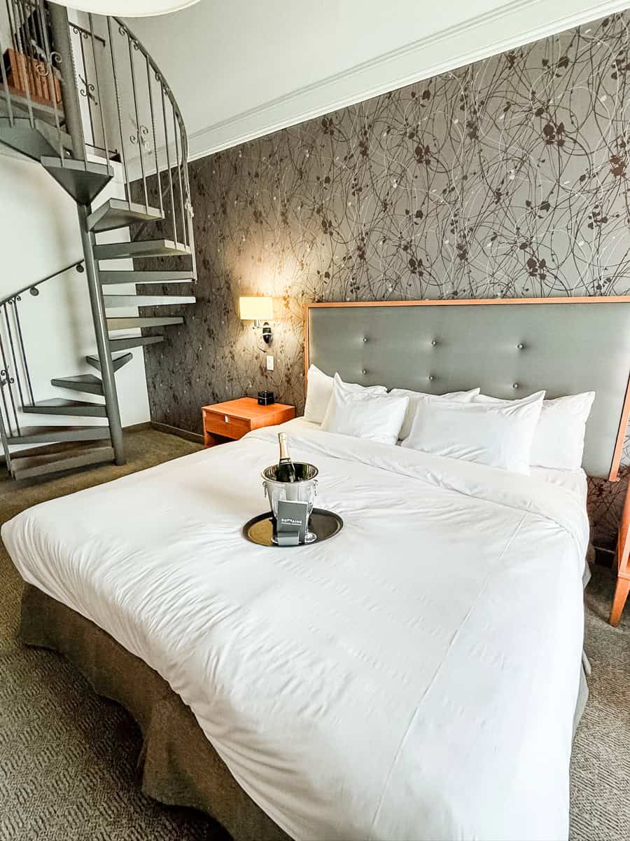 beautiful hotel room with king bad, fine-like wallpaper, and a spiral staircase up to the next level. On the bed is a bottle of sparkling wine as a welcome gift