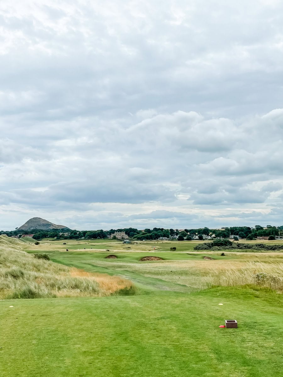 Expansive view of a coastal links-style golf course with rolling fairways, tall fescue grass, and several deep bunkers under a cloudy sky, with a distant hill and seaside town visible in the background.