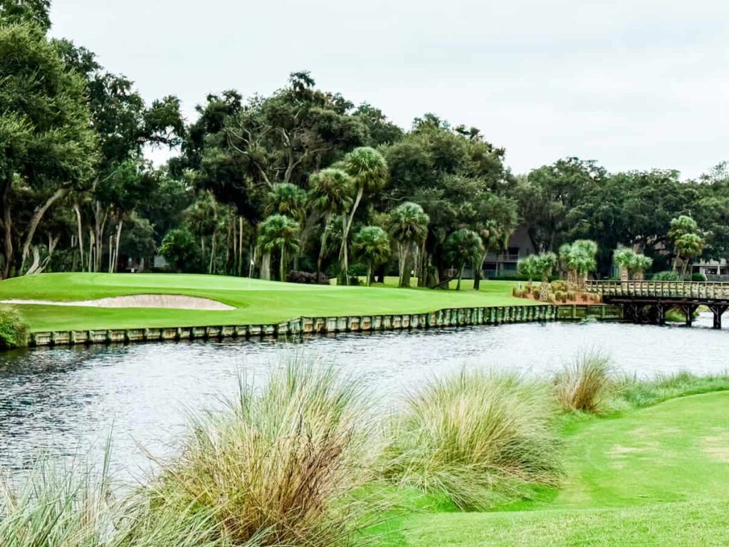 Scenic view of a Hilton Head golf course featuring a lush green fairway bordered by palm trees, a wooden bridge crossing over calm water, and a sand bunker beside the putting green under a cloudy sky.
