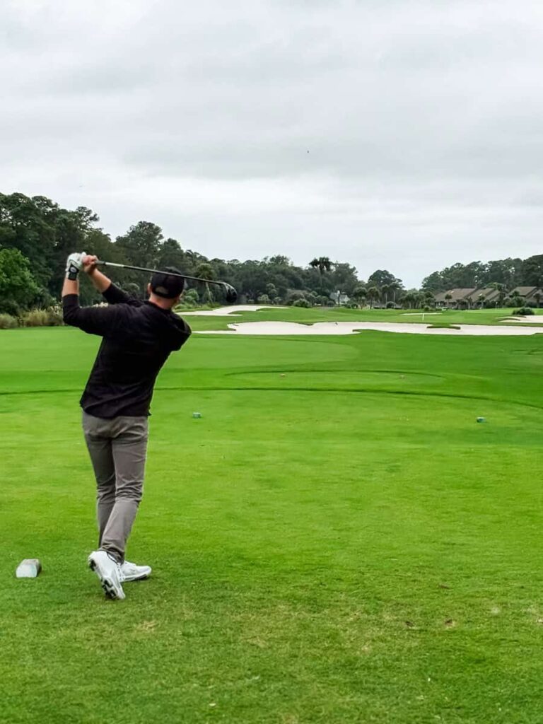A golfer follows through on a tee shot under cloudy skies at a lush, well-manicured golf course. The view shows the fairway framed by sand bunkers and trees in the distance, with homes bordering the course on the right side.