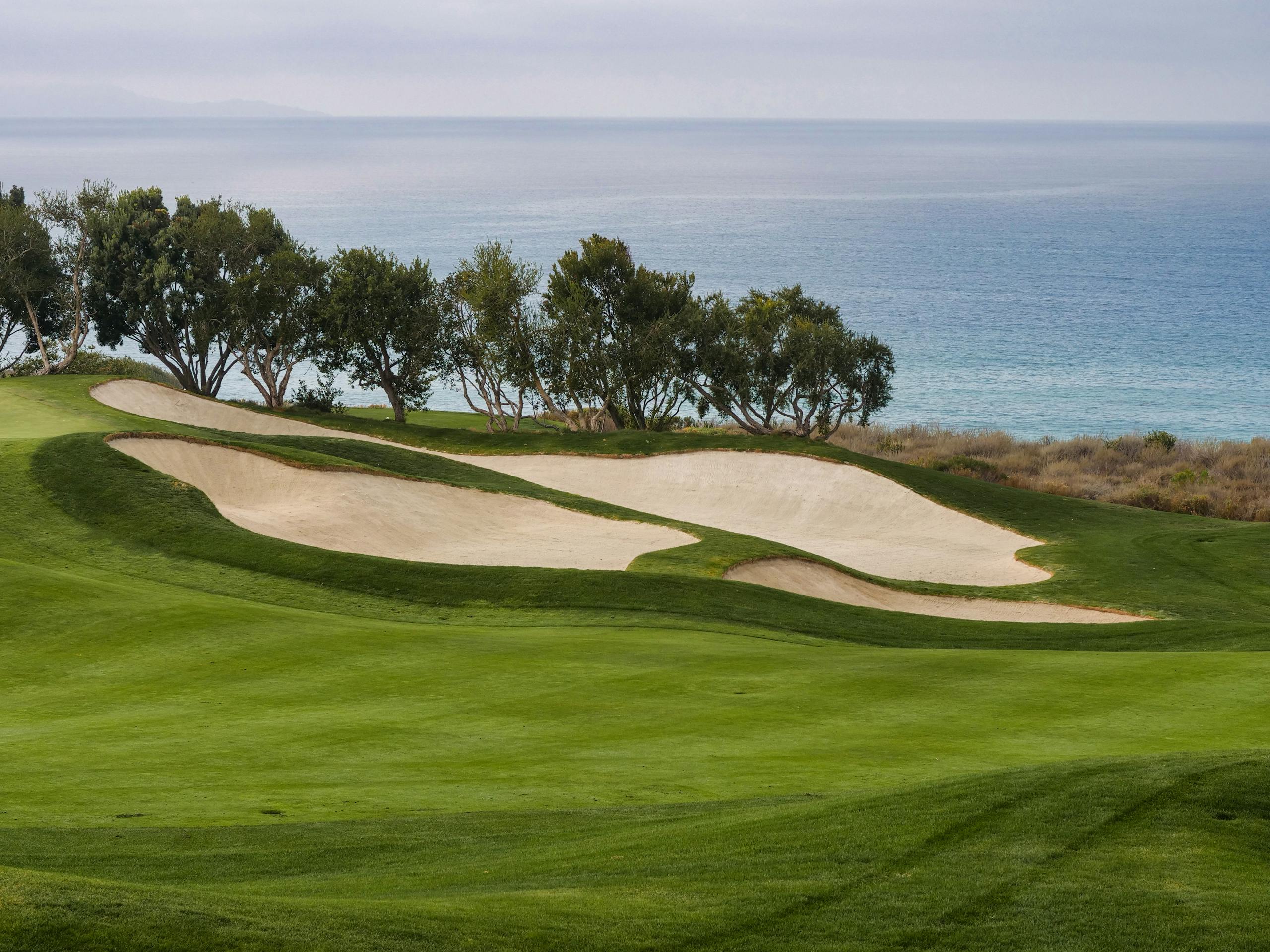 Golf course by the sea with green fairway and sandy bunkers under a cloudy sky.
